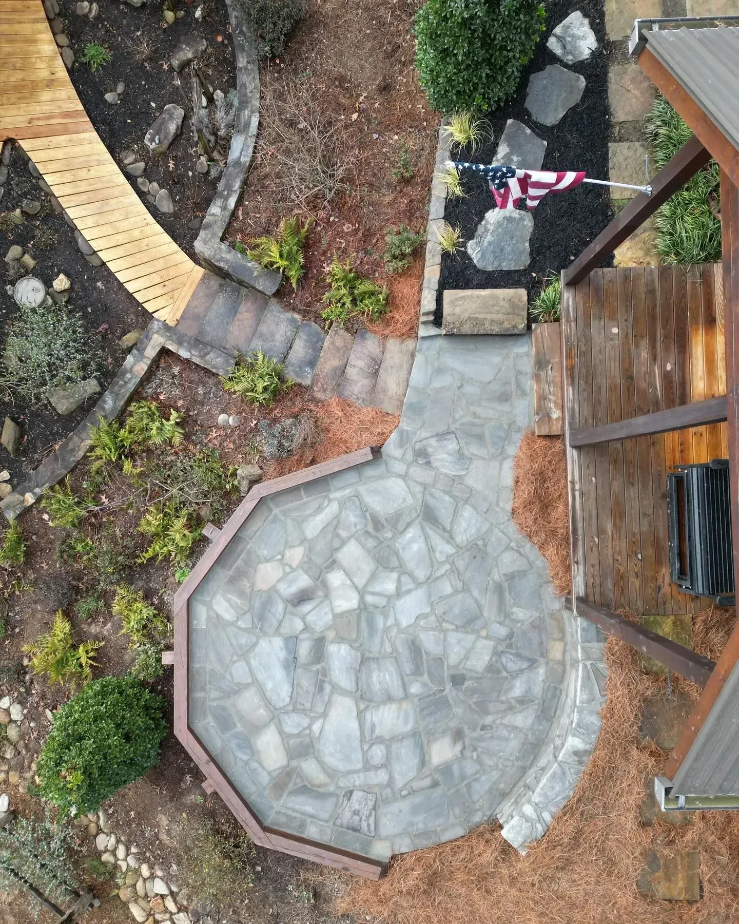 Top-down view of a stone patio and garden path beside a wooden deck and a striped barrier tape.