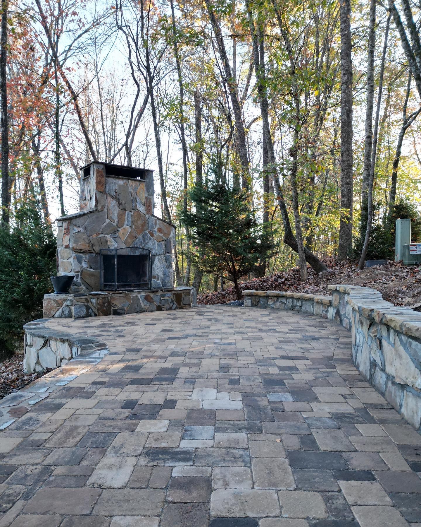 Stone outdoor fireplace beside a paved patio, surrounded by trees in a wooded backyard.