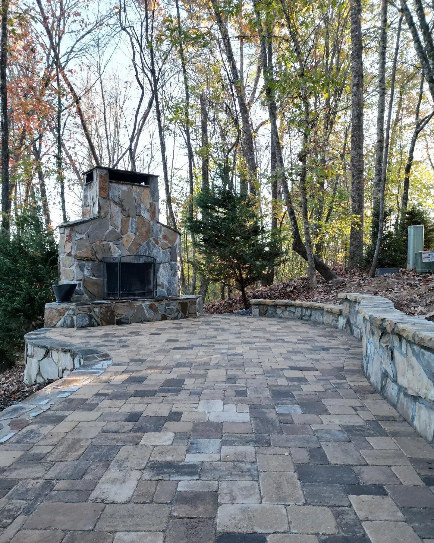 Stone patio with a large outdoor fireplace, flanked by low walls and surrounded by trees in autumn.