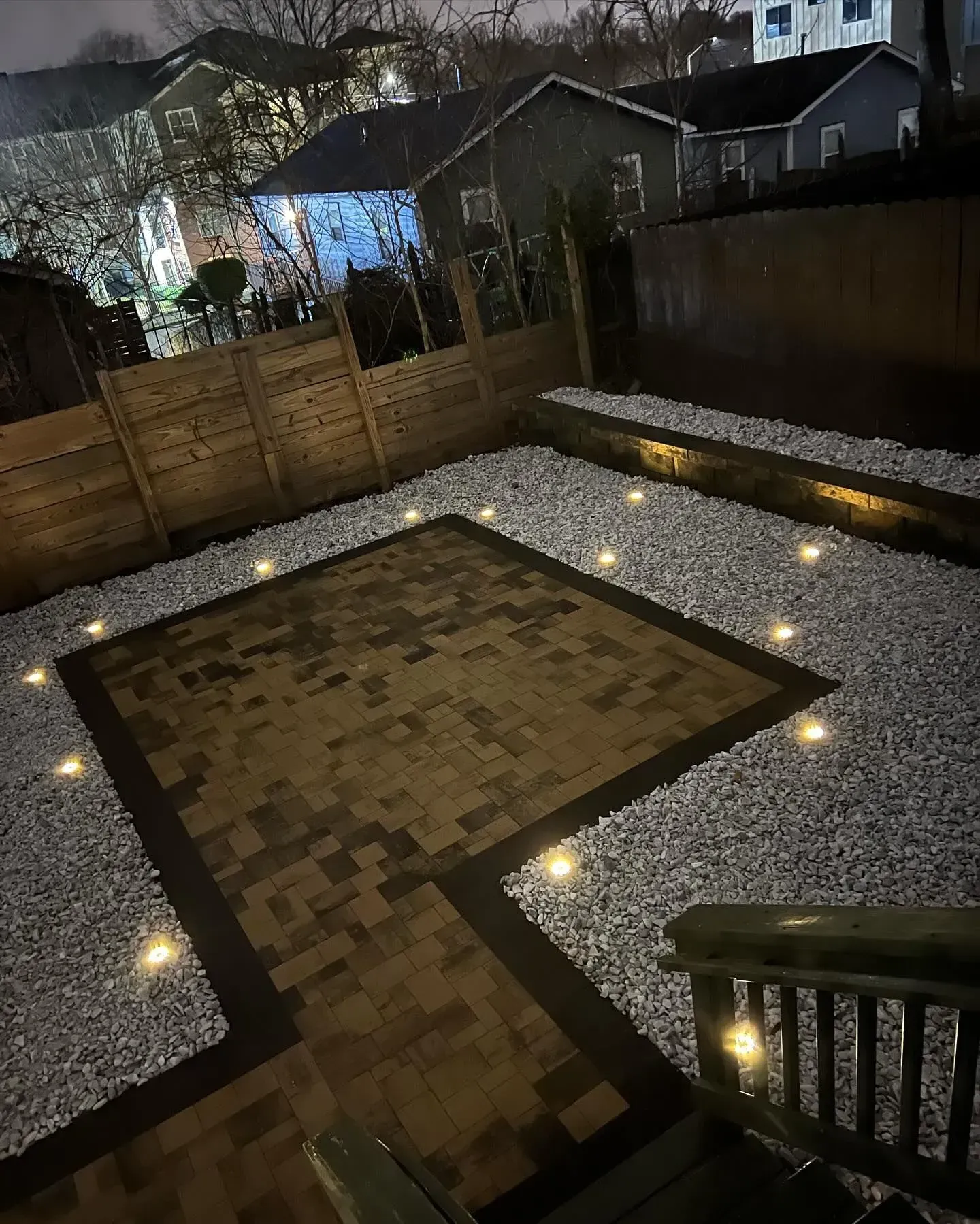 Nighttime backyard patio with a paved square, gravel border, fence, and small ground lights