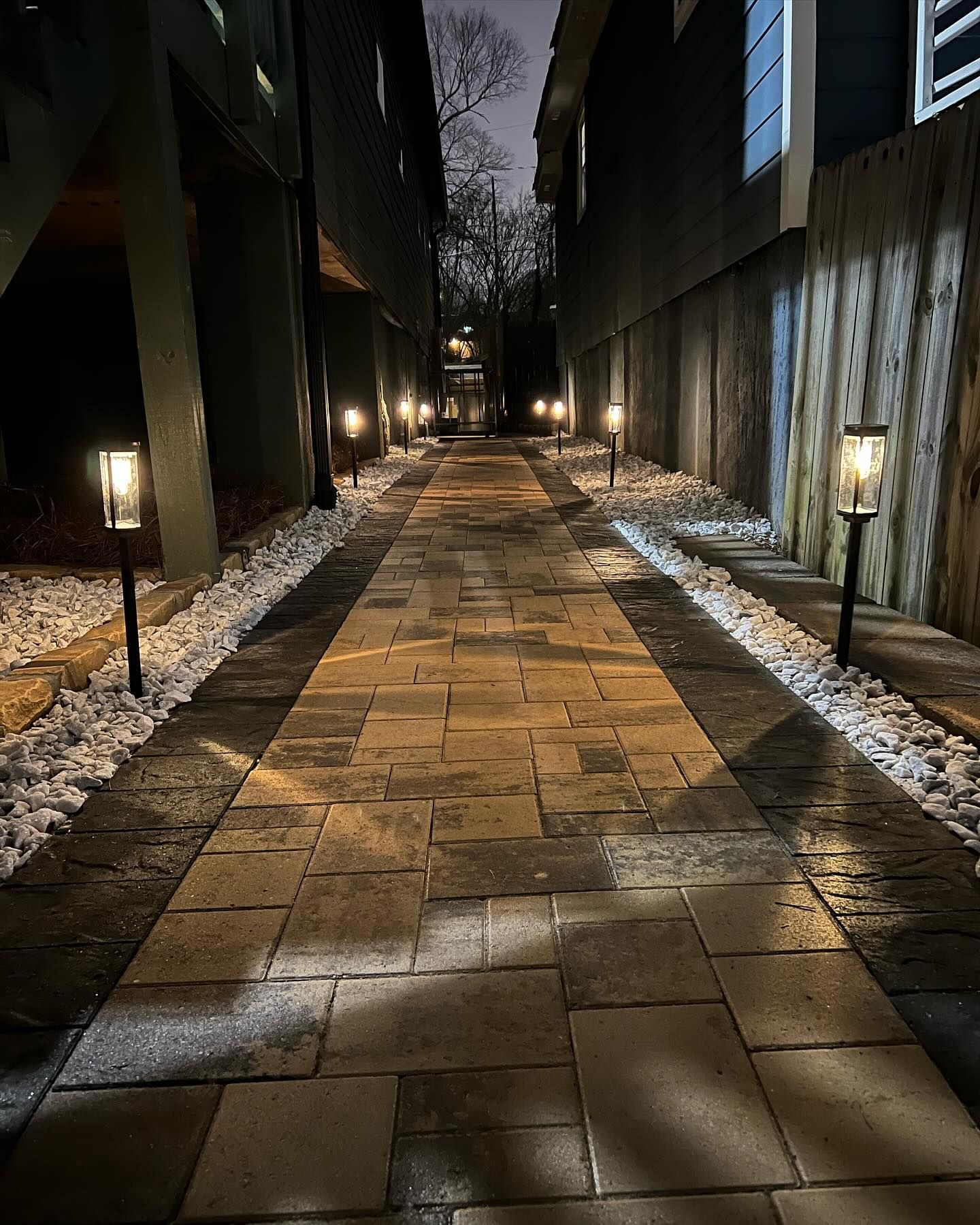 Nighttime stone walkway lit by lanterns between tall buildings, with snow along the edges.