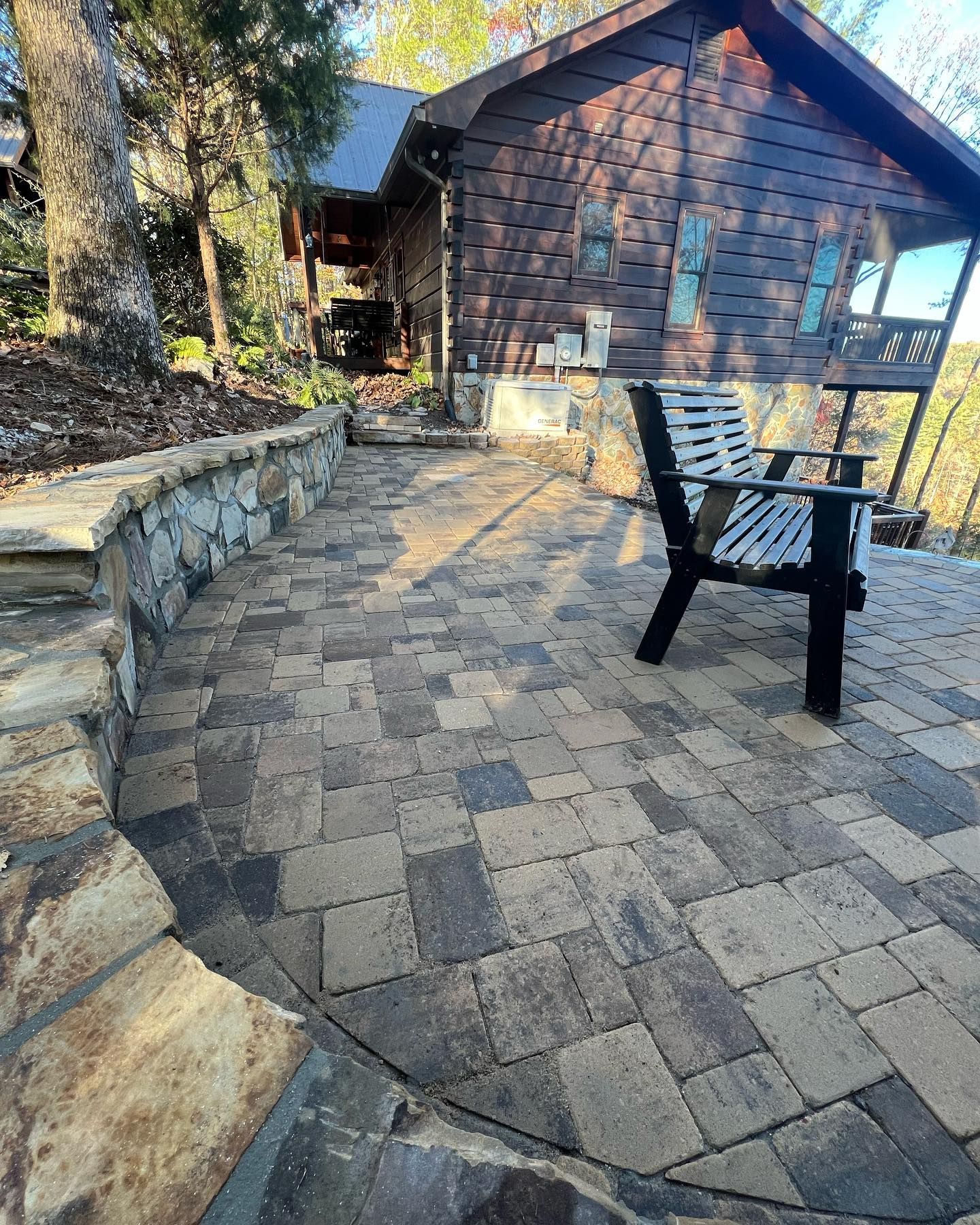 Stone patio with a black chair beside a rustic cabin in a wooded yard.