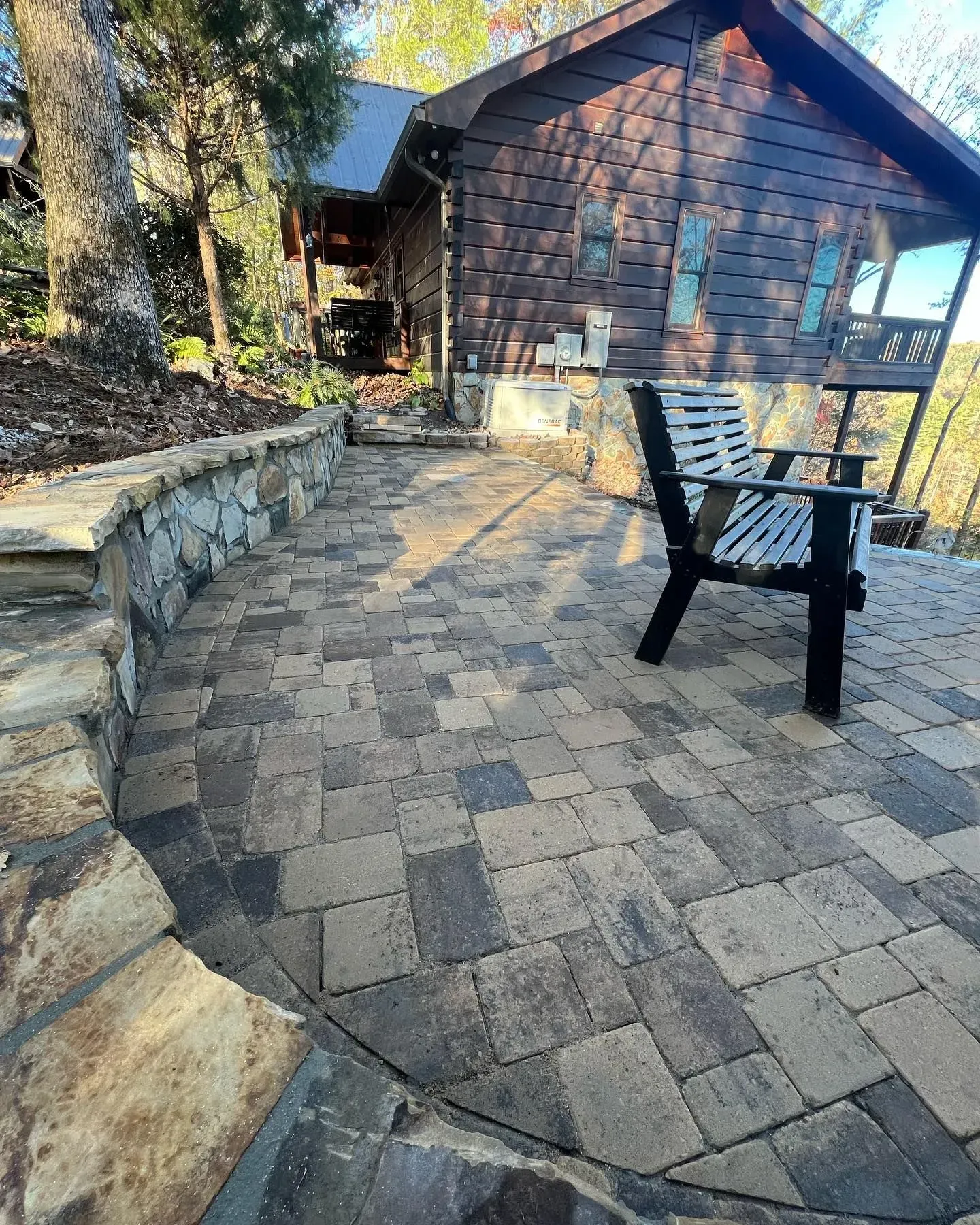 Stone patio with black bench beside a rustic cabin and stone steps in a wooded setting