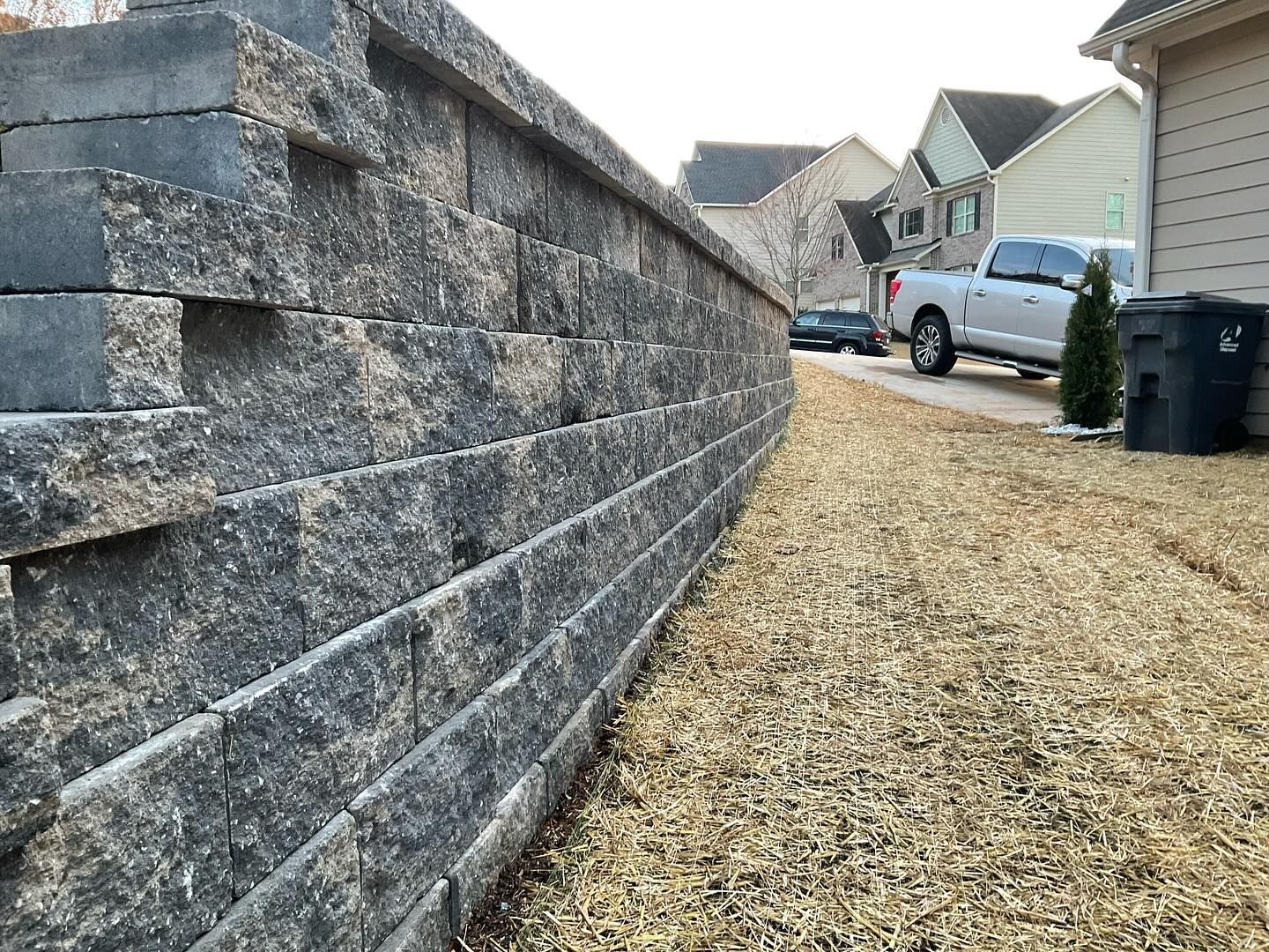 Stone retaining wall along a gravel driveway beside suburban houses and parked cars