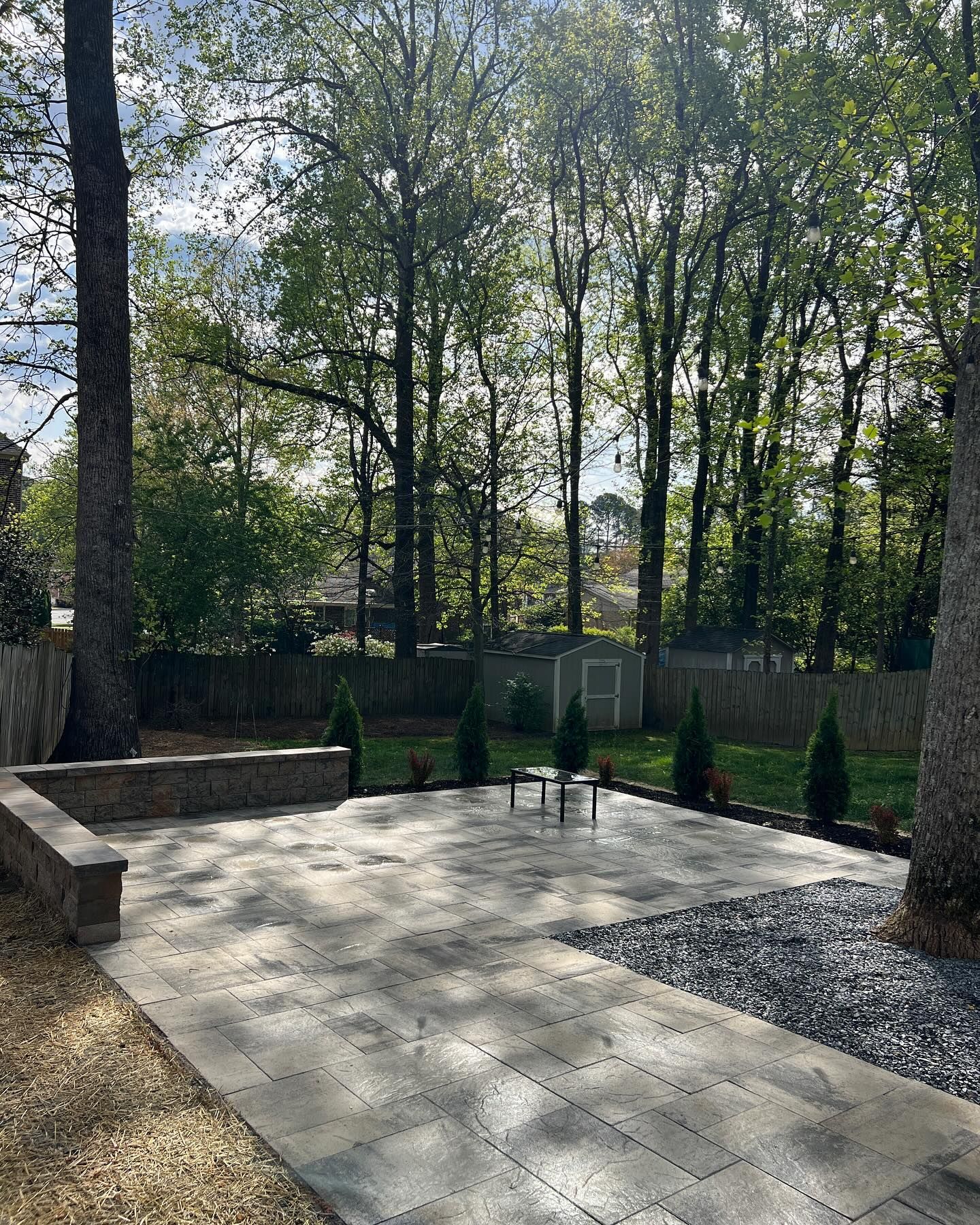 Backyard patio with stone pavers, gravel border, and tall trees in sunlight