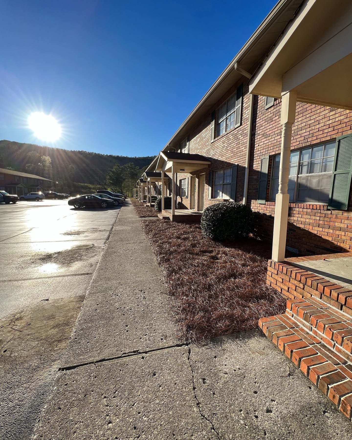 Sunny parking lot beside a brick apartment building with covered walkway and shrubs