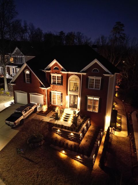 Illuminated brick house at night with lit windows, driveway, and warm pathway lights in front yard
