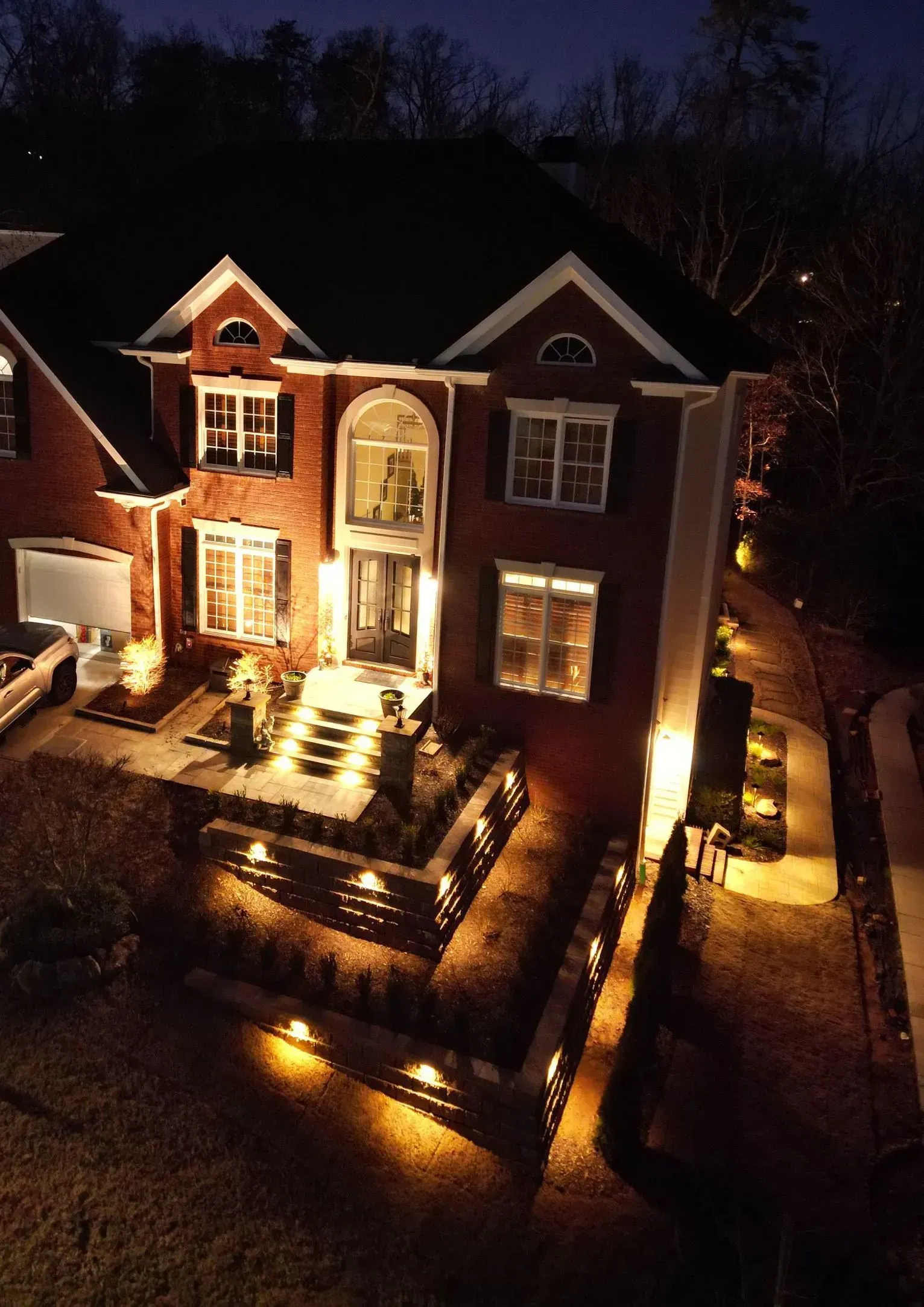 Large brick house lit by warm exterior lights at night, with illuminated walkway and steps