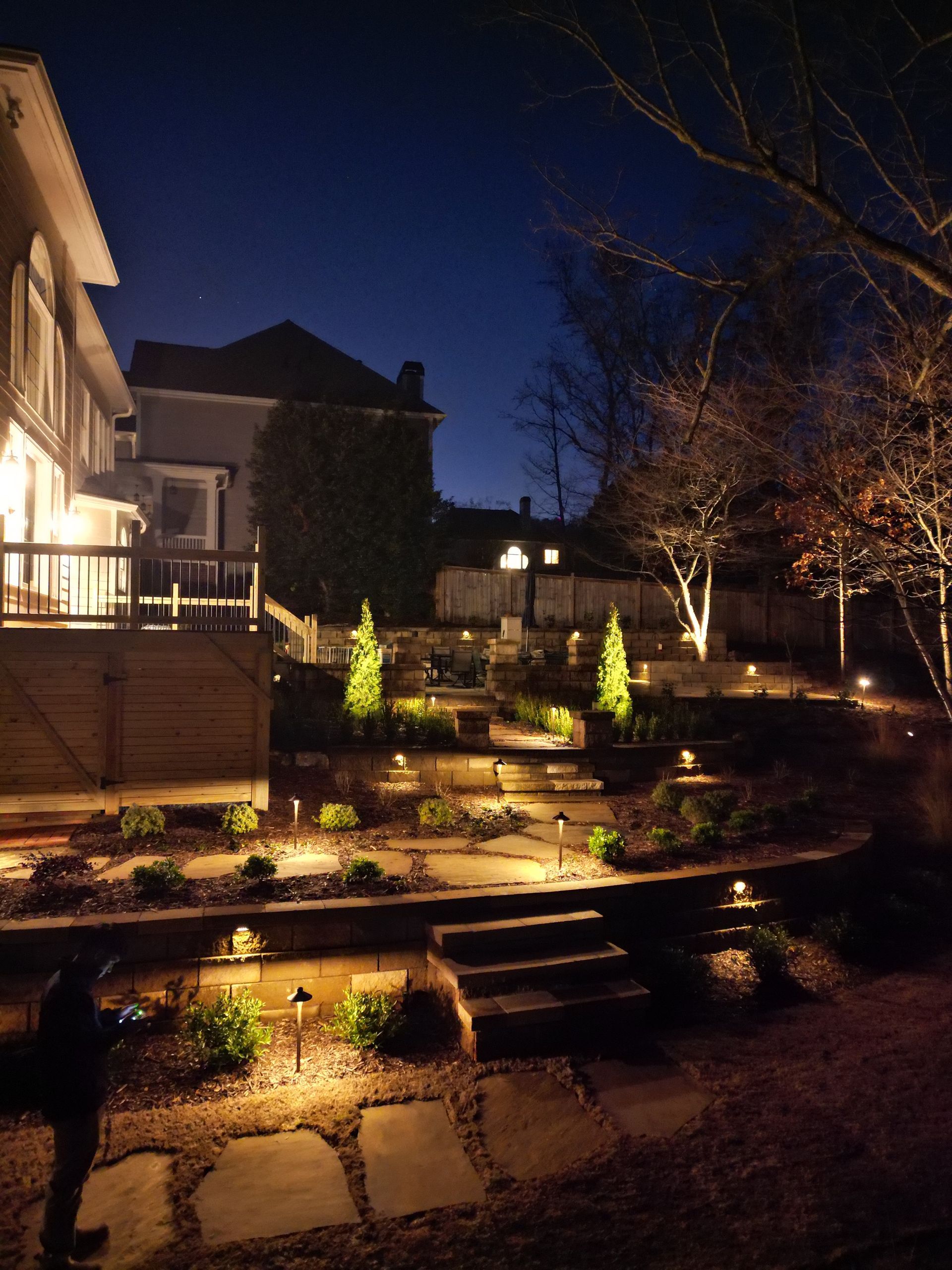 Lit backyard terrace at night with stone steps, landscaped beds, and warm house lights
