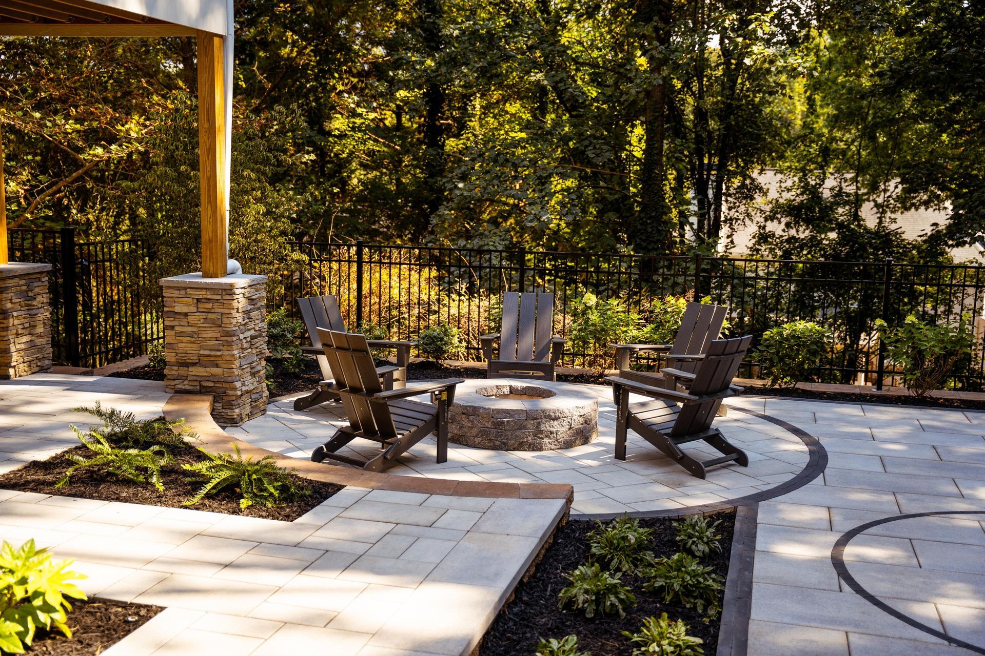 Outdoor patio with two wooden chairs around a stone fire pit, set on a paved courtyard with trees and fence.