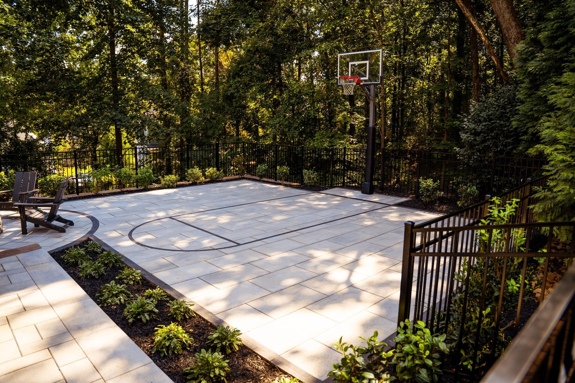 Outdoor basketball half-court surrounded by trees and a black fence in a wooded backyard