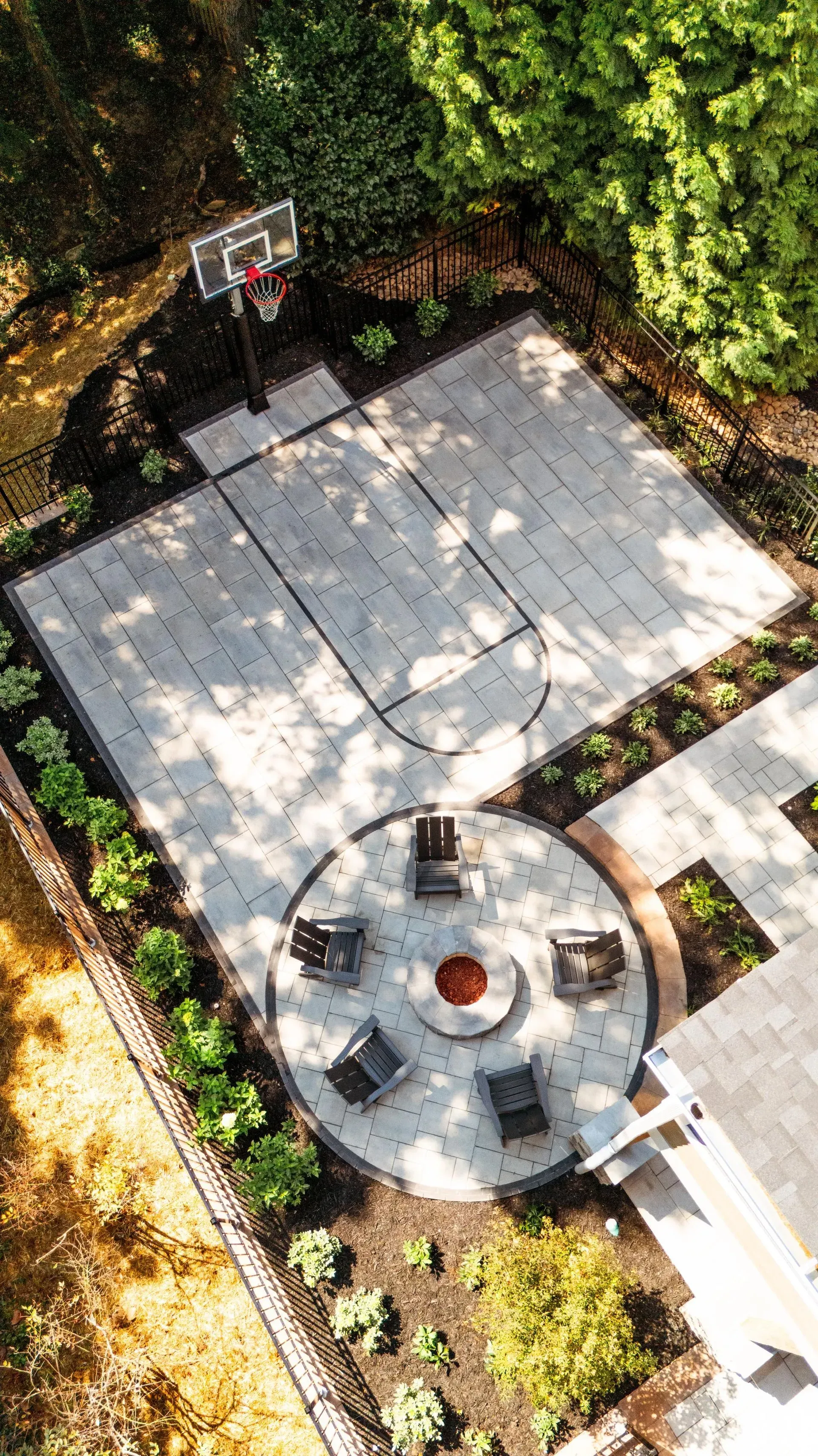 Aerial view of a backyard patio with a round fire pit, paved seating area, and basketball hoop, bordered by trees.