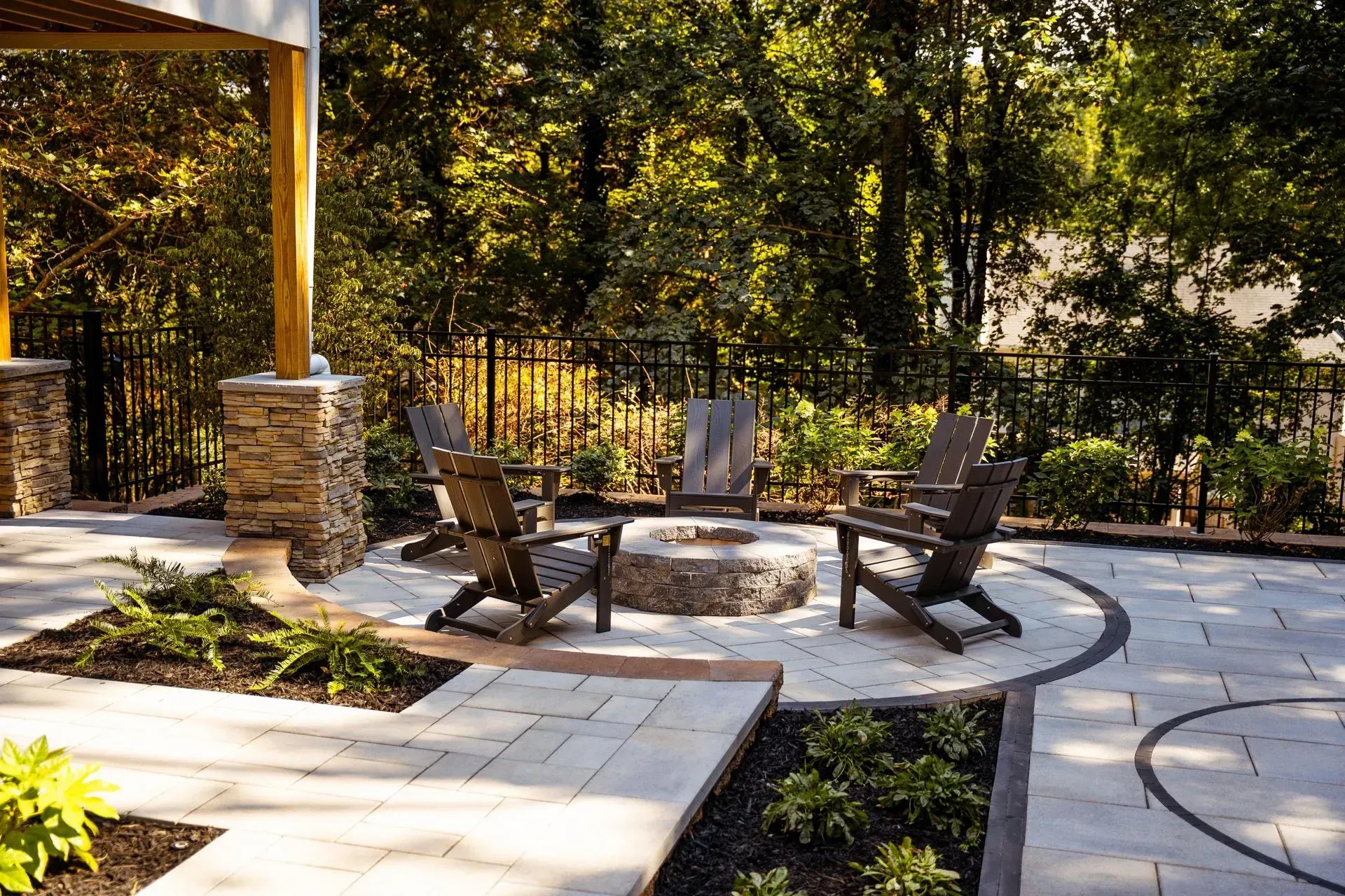 Outdoor patio with stone fire pit, wooden chairs, and shaded trees in a landscaped garden