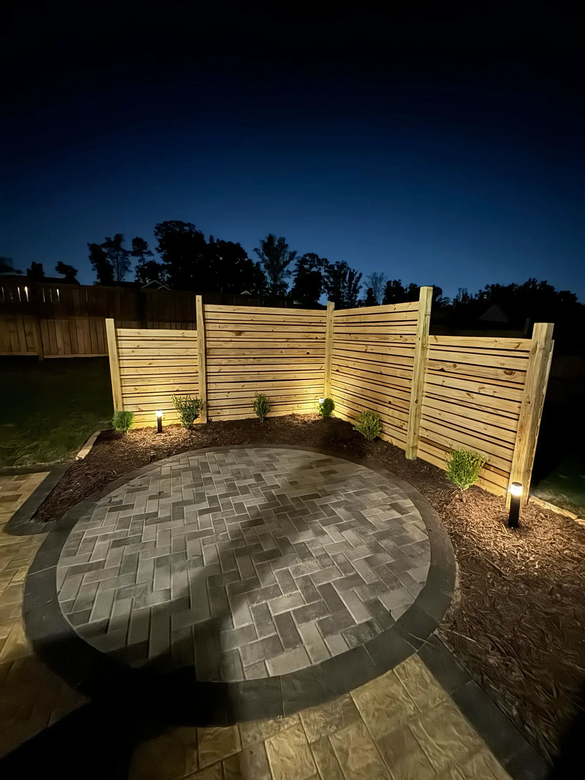 Nighttime backyard patio with circular stone fire pit, wooden privacy fence, and pathway lights