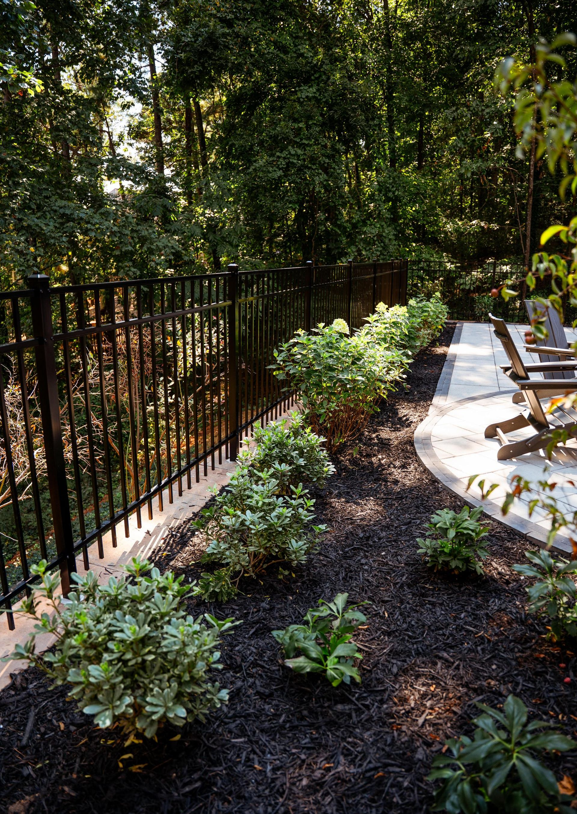 Shaded garden path with black fence, lush greenery, and a curved stone patio with chairs