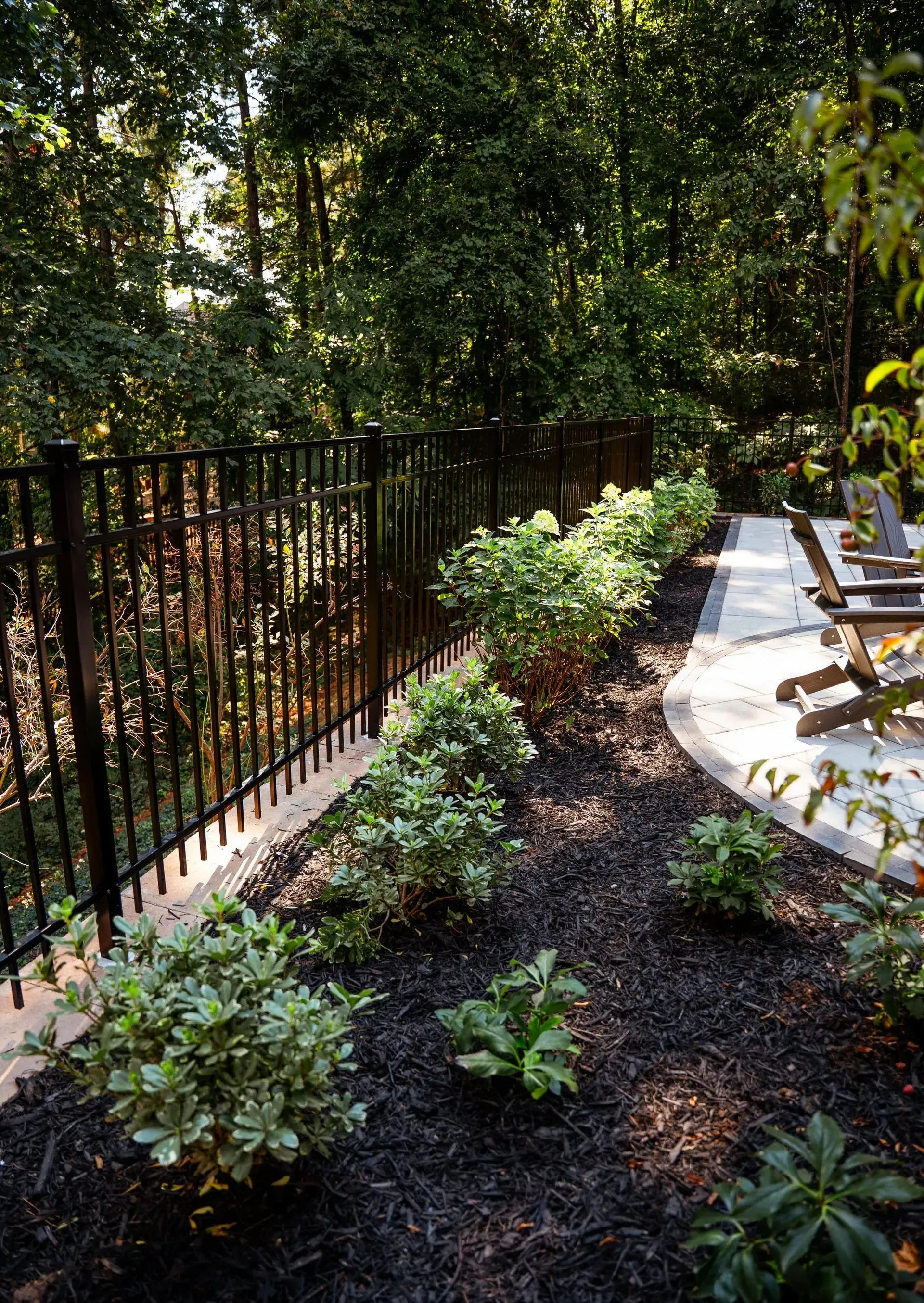 Garden path beside a black fence, with lush plants and patio chairs in dappled sunlight.