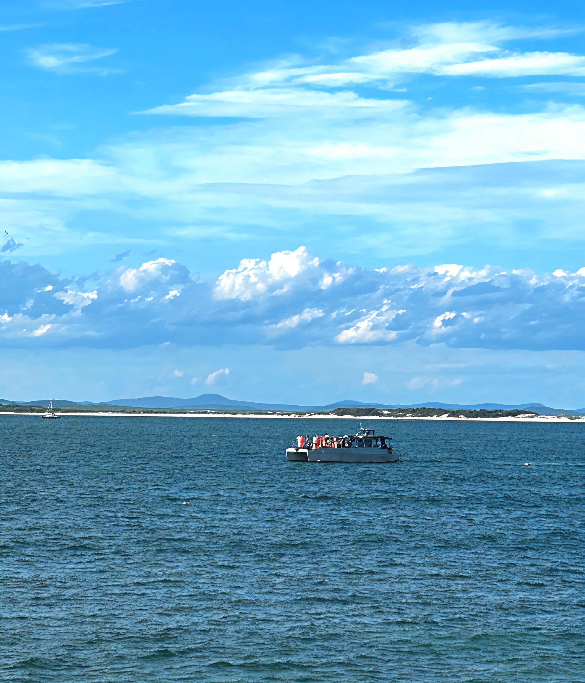 Boat sailing on calm blue water under a partly cloudy sky, a distant sandy shoreline.