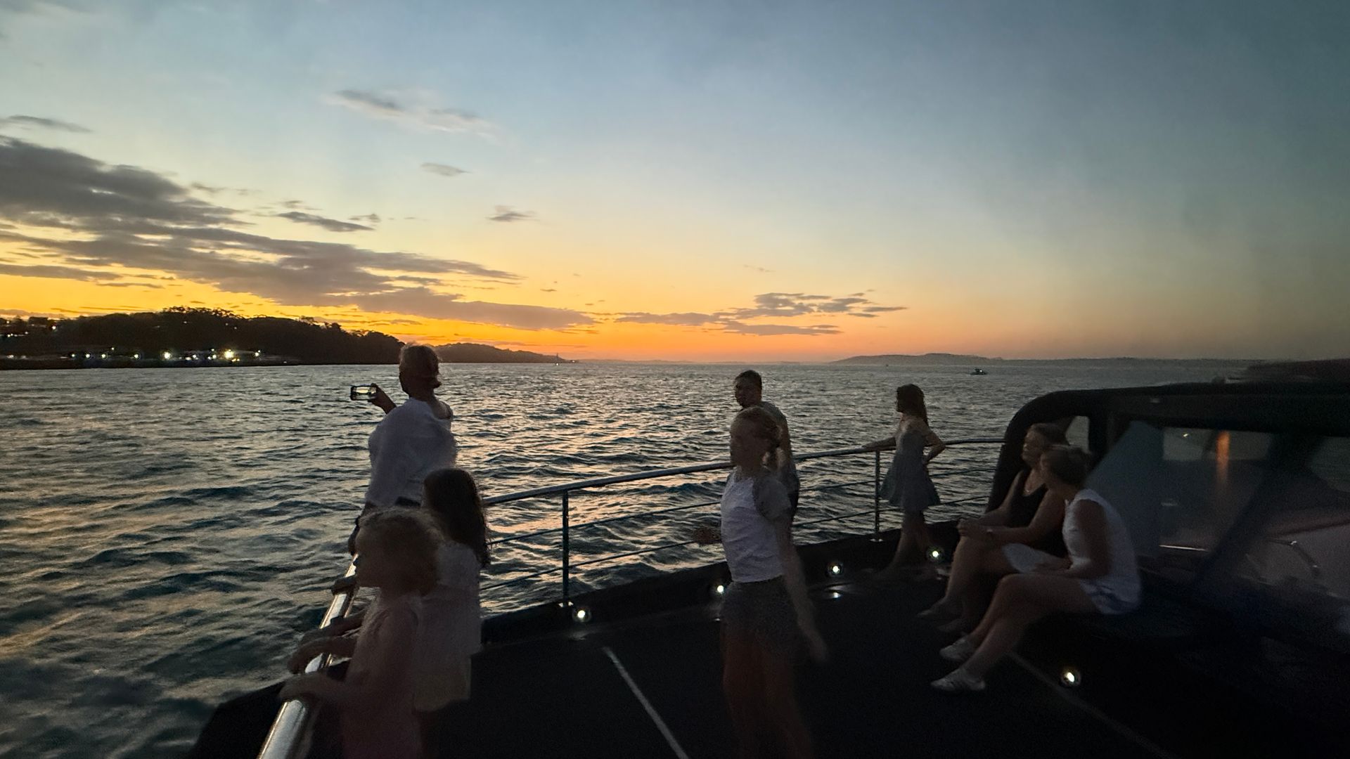 People on a boat watch the sunset over the water; orange and yellow sky, dark silhouettes.