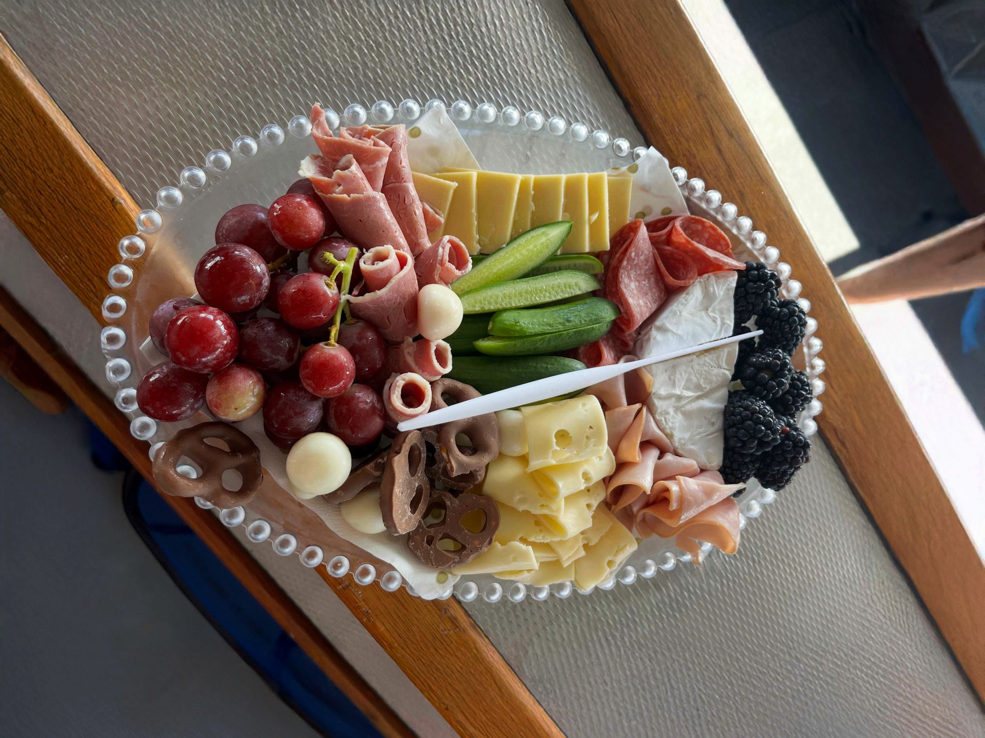 A charcuterie board featuring grapes, deli meats, two types of cheese, sliced cucumbers, blackberries, and chocolate.