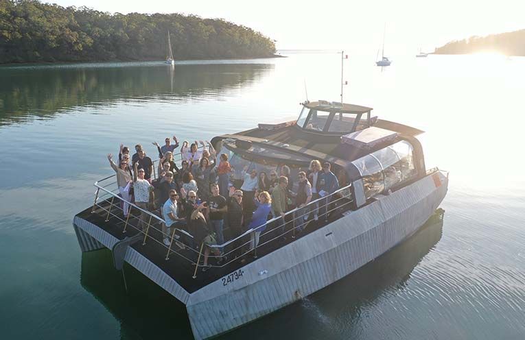 An aerial view of a boat crowded with a group of people waving while cruising on calm, sunlit water near a wooded shore.