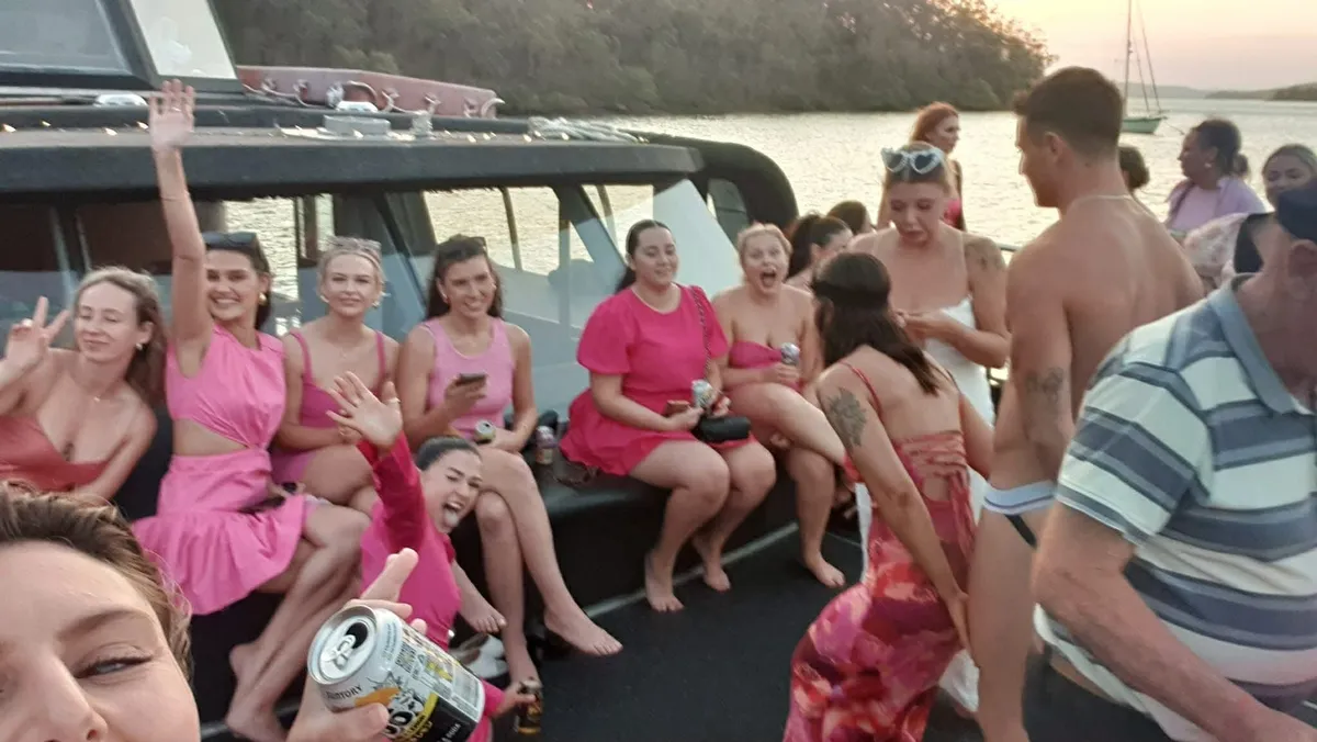 A group of people wearing pink clothes celebrate together on the deck of a boat at sunset, with some waving and smiling.