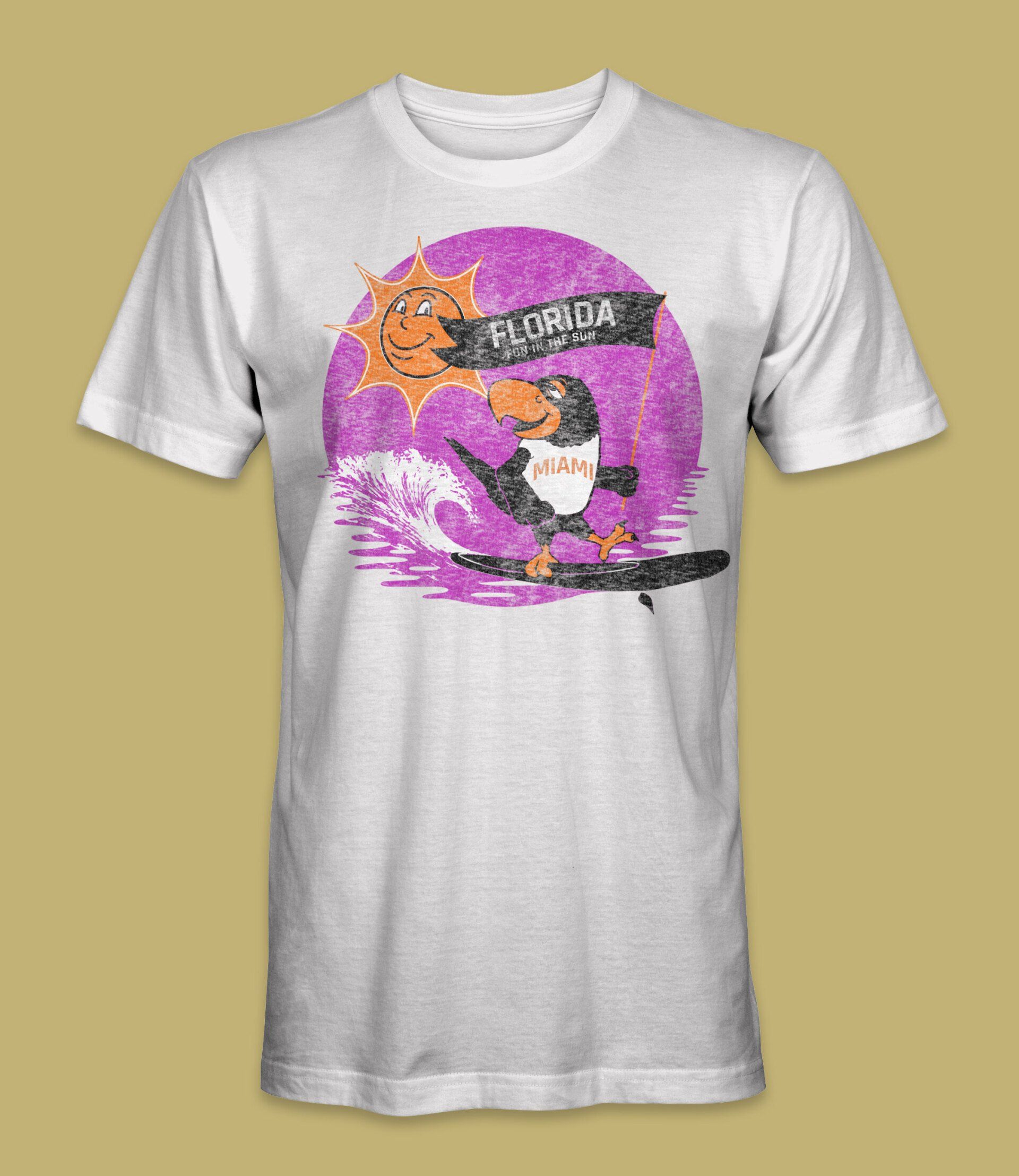 White t-shirt with a surfing penguin illustration. Pink sun, purple wave, and 