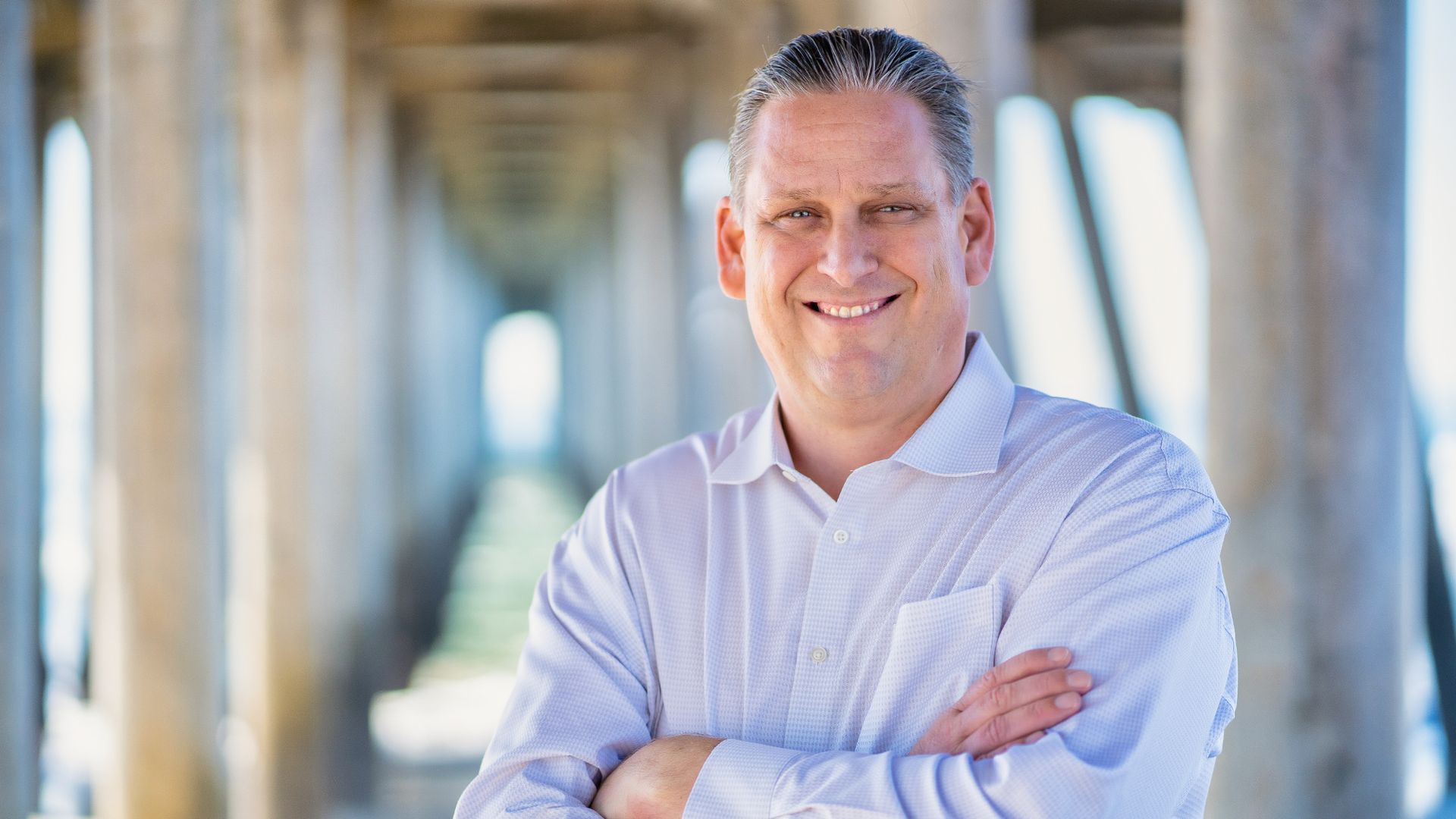 A man in a white shirt is standing under a pier with his arms crossed . and smiling. Tony Strickland