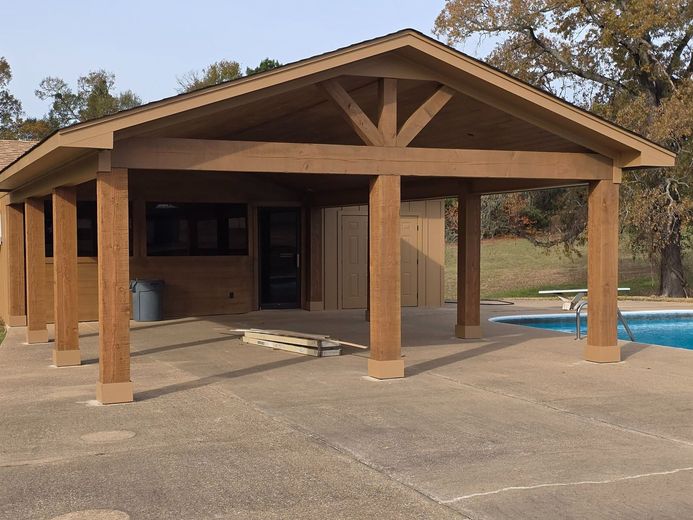 Wooden pavilion with a gabled roof and open sides beside a swimming pool in a park setting
