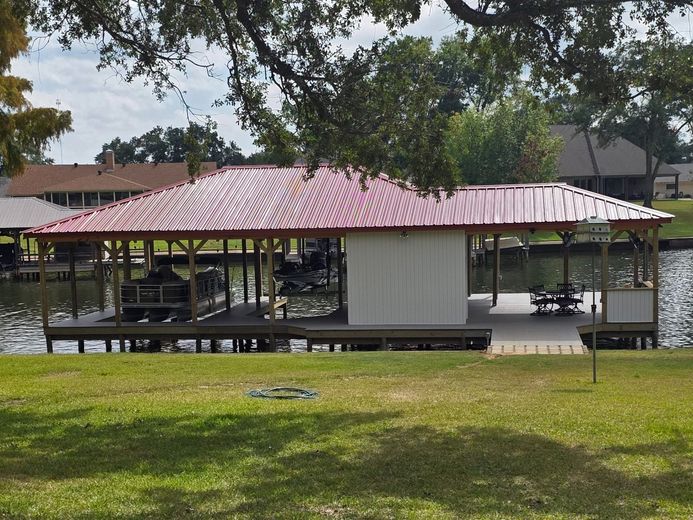 Open-air pavilion with red roof in a grassy park, surrounded by trees and a paved walkway.