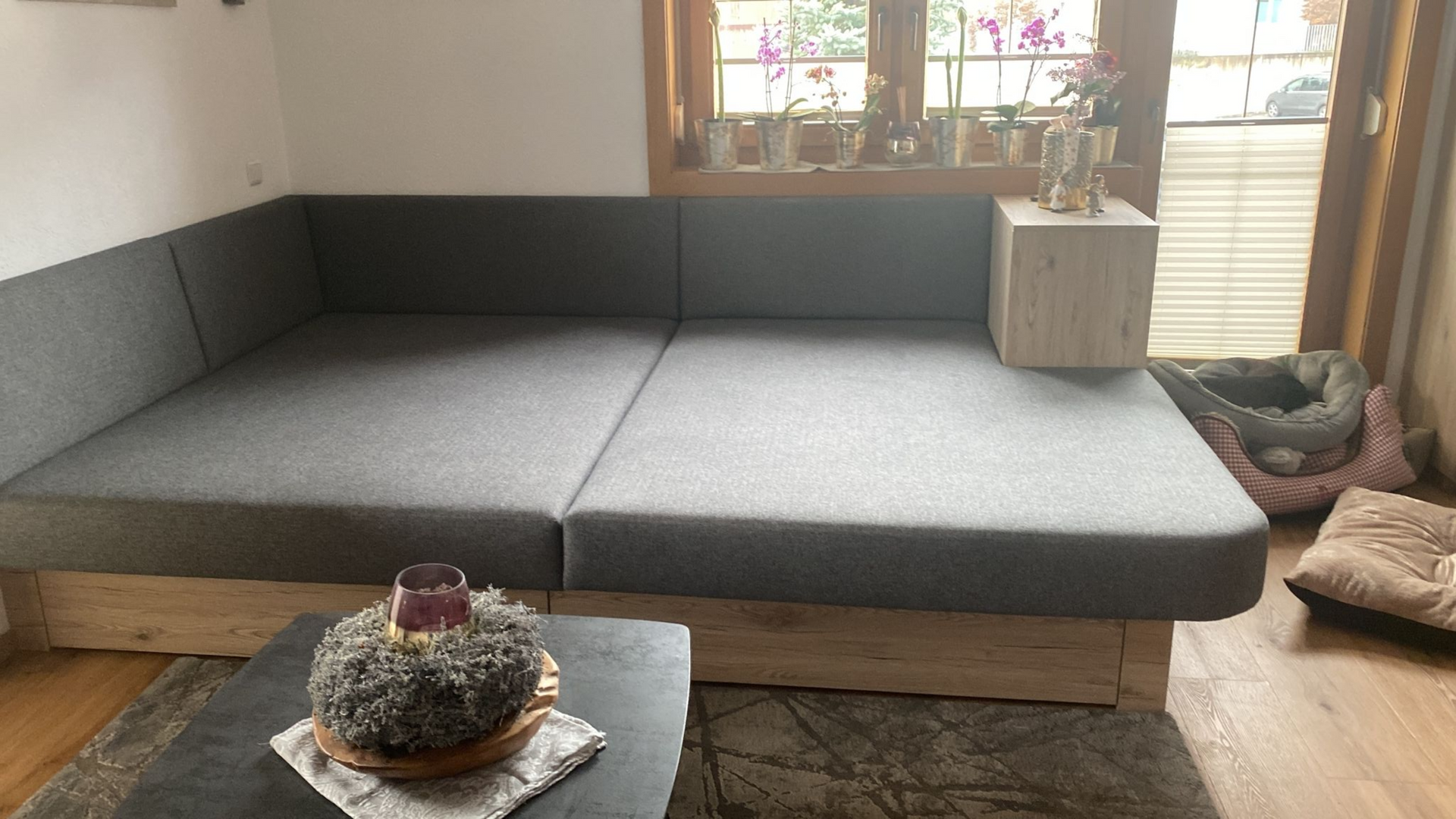 In einem Zimmer mit Fensterbank, Pflanzen und einem kleinen Hundebett steht ein graues Ecksofa mit hellem Holzgestell.