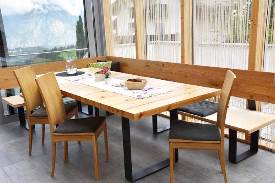 Ein Esstisch mit Stühlen und einer hölzernen Eckbank in einem hellen, modernen Zimmer mit großen Fenstern und Bergblick.