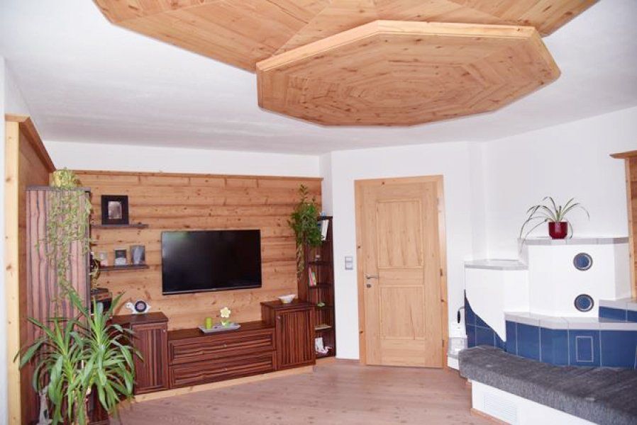 Ein rustikales Wohnzimmer mit holzgetäfelter Wand, Flachbildfernseher, Holzdecke, Tür und einem blauen Kachelofen.