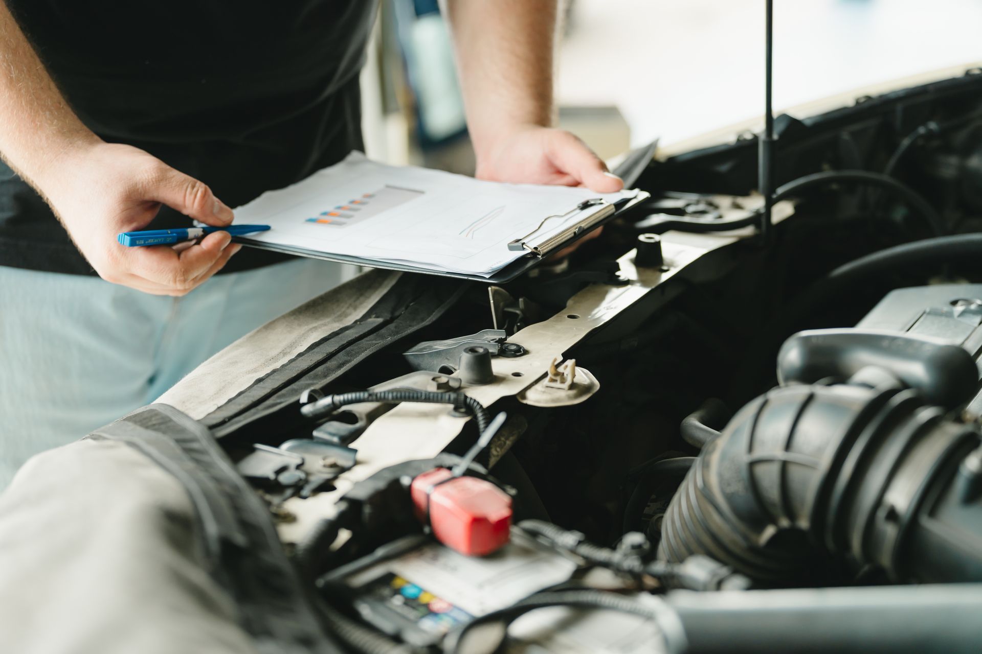 Person holding a clipboard and pen beside an open car engine in a repair shop