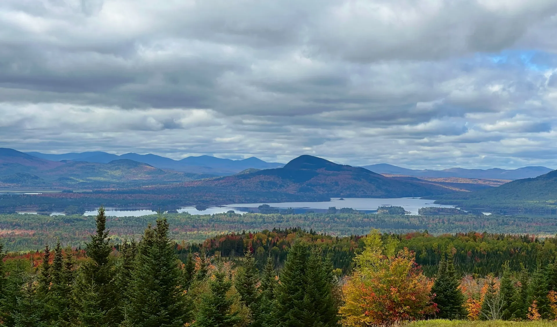 Cloudy mountain landscape with a lake, distant town, and autumn-colored trees in the foreground
