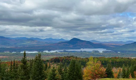 Cloudy mountain landscape with a lake, distant town, and autumn-colored trees in the foreground