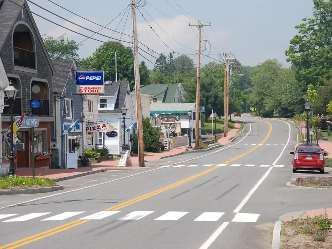Quiet curving road with a red car, small roadside buildings, and trees on a sunny day