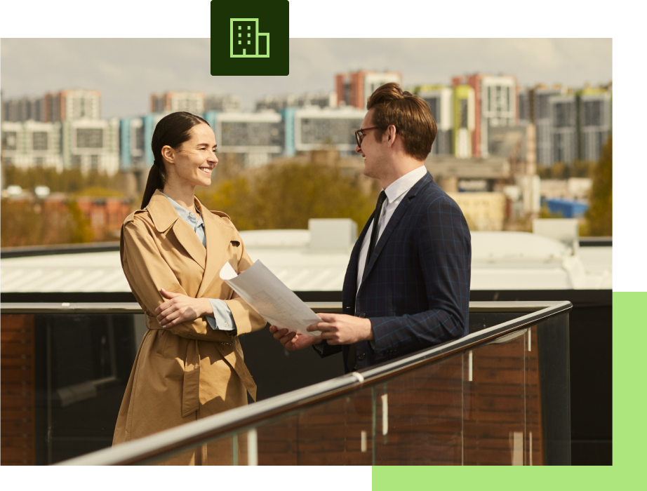 Two people on a rooftop, looking at documents, with buildings in the background.