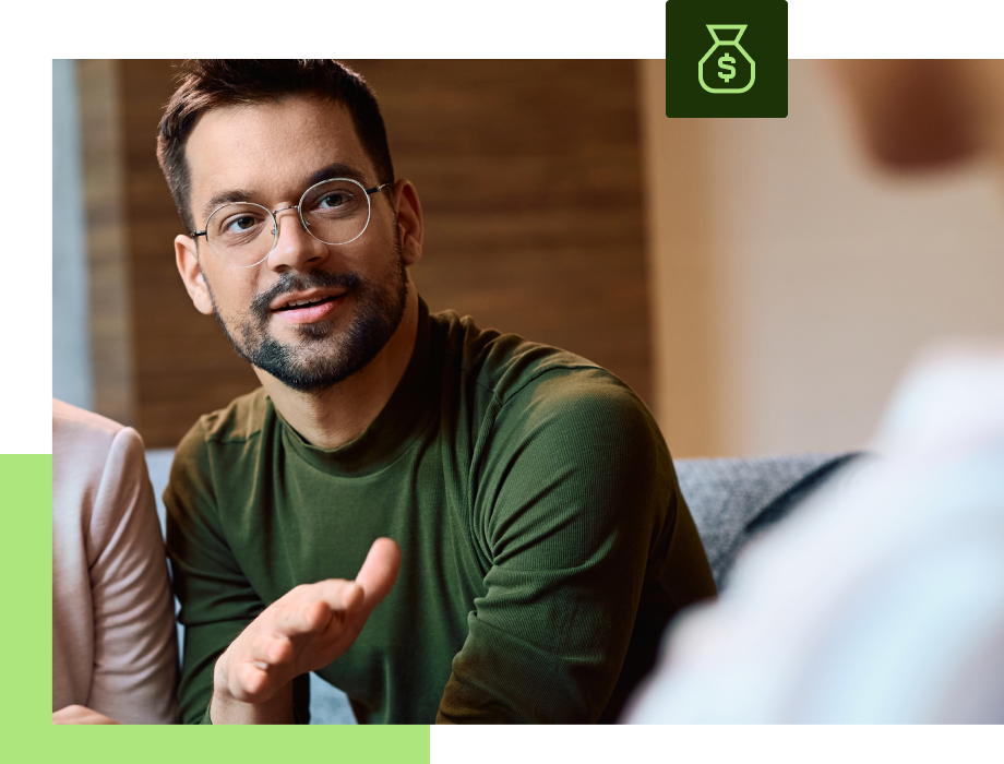 Man in glasses gesturing, speaking in a meeting. Green shirt. Money bag icon in top right.