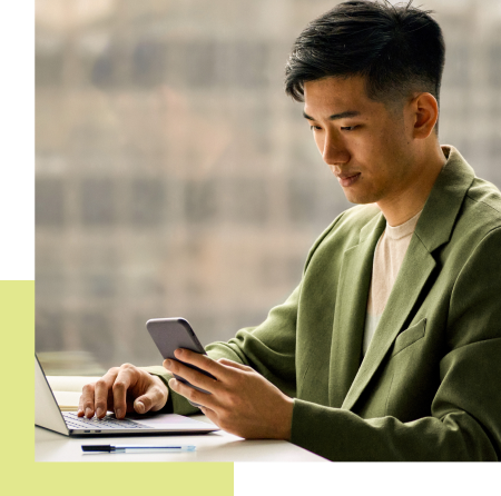 Man in green blazer using a laptop and phone, looking focused near a window.