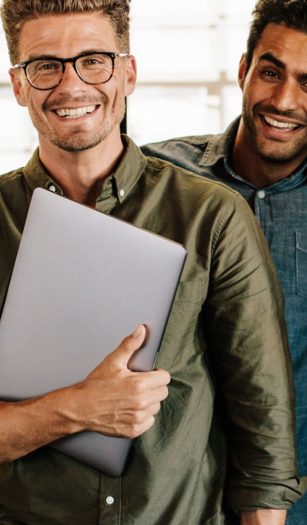 Two men smiling at camera; one holds a laptop.