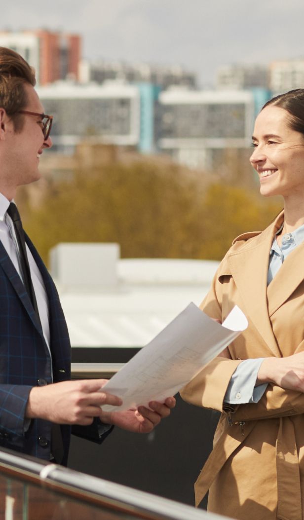 Two people, smiling, on a rooftop; one holds papers.