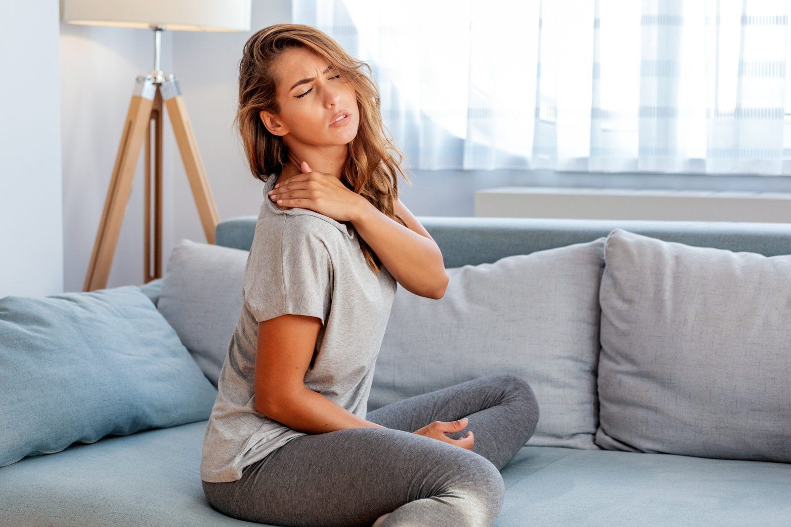 Woman sitting on a couch, holding her neck in pain. Indoor setting, soft colors.