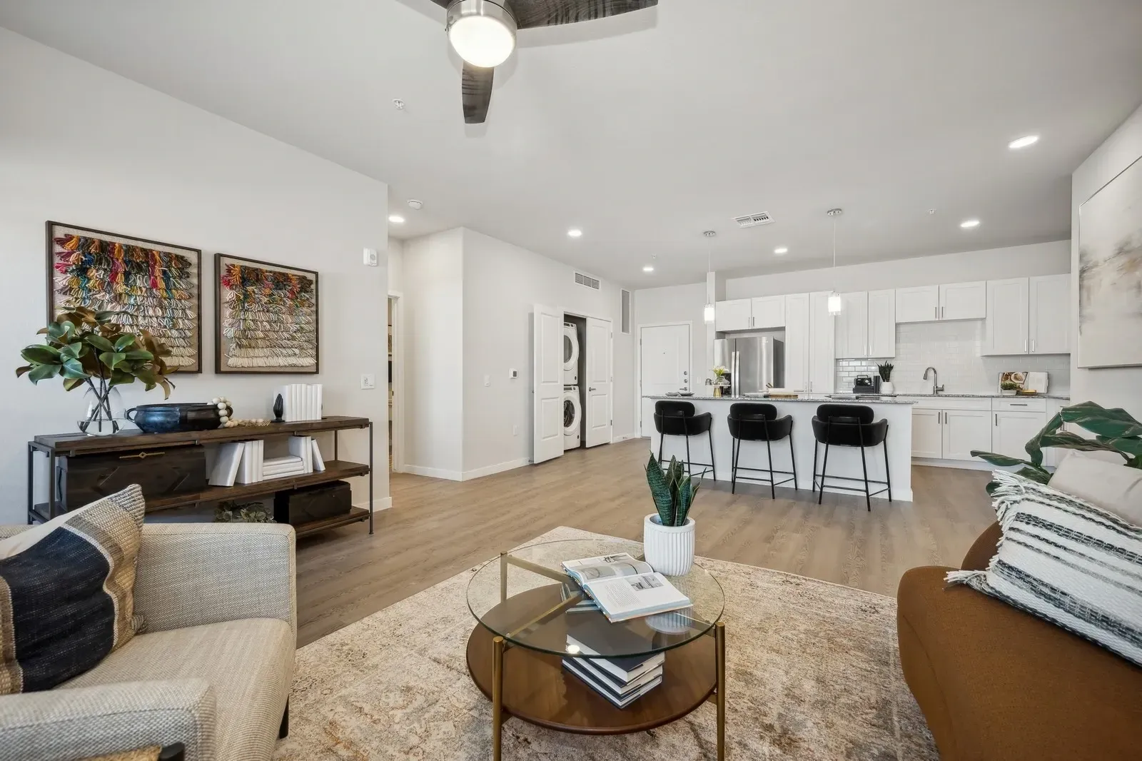 Living room and kitchen area with white cabinets, island with stools, and stackable washer and dryer at Longbow Luxury apartments in Mesa, AZ.