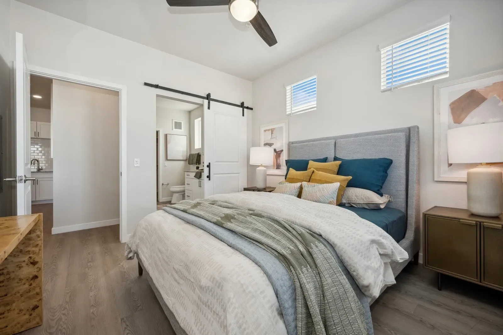Modern bedroom with a king-size bed, barn door leading to bathroom, and view of kitchen at Longbow Luxury Apartments Mesa, AZ.