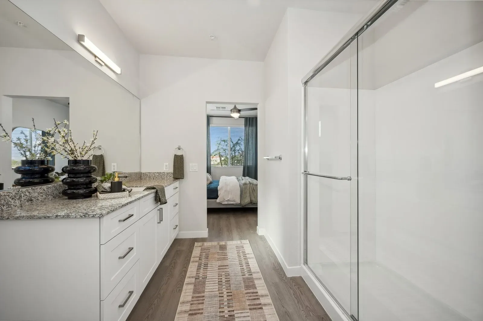 Modern bathroom with double vanity, granite countertops, and glass-enclosed shower. View into bedroom.
