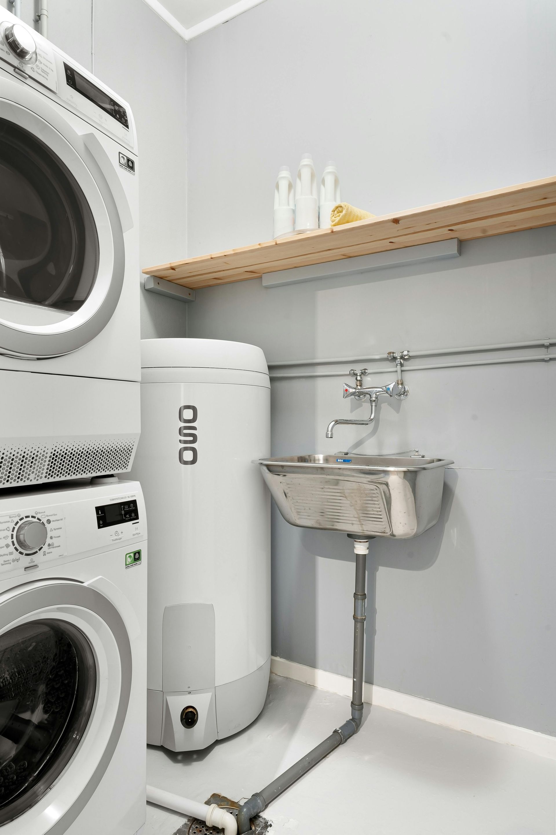 Cuarto de lavandería con lavadora y secadora apiladas, fregadero, calentador de agua y estante con botellas.