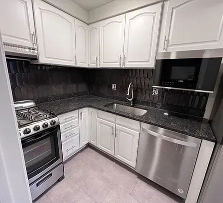 Small, white kitchen with stainless steel appliances and dark countertops and backsplash.
