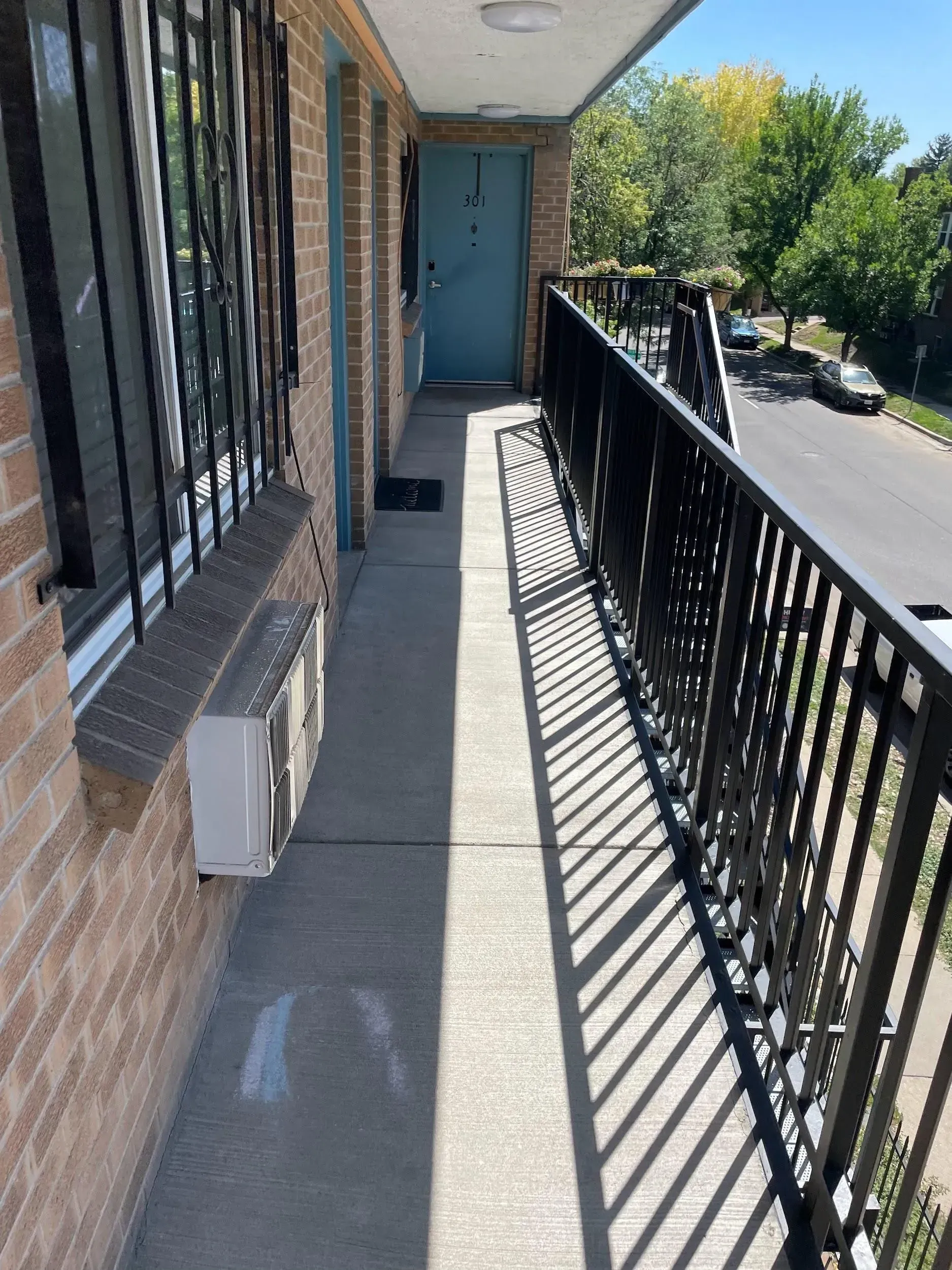 Narrow apartment balcony with brick wall, metal railing, and a blue door.