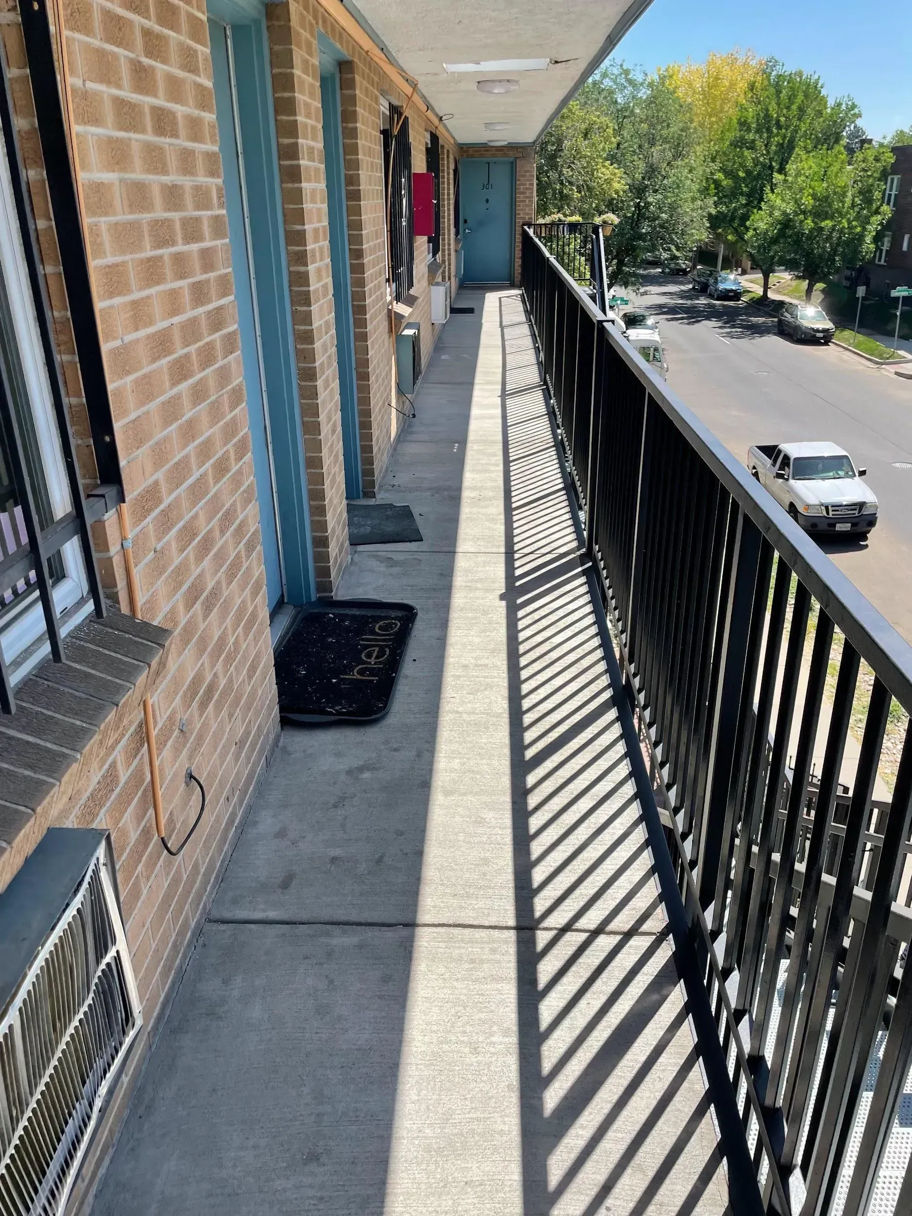 Exterior apartment balcony with black railing, brick wall, and blue door. Cars visible on street.