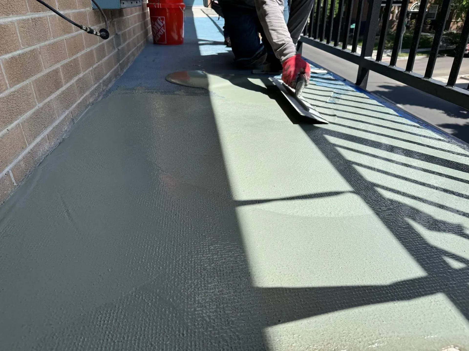 Person smoothing light-colored concrete on a balcony with a trowel. Brick wall, black railing, sunny day.